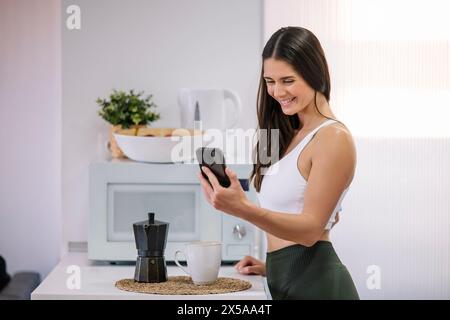 Una donna aderente all'abbigliamento sportivo sorride mentre usa il suo smartphone in una cucina moderna Foto Stock