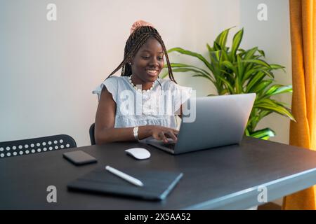 Un'allegra donna afroamericana lavora con attenzione da casa, coinvolgendo il suo notebook in un ambiente confortevole e luminoso Foto Stock