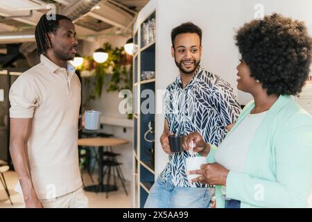 Tre professionisti impegnati in una conversazione informale in un moderno spazio di coworking tenendo in mano tazze di caffè Foto Stock