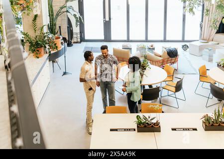 Un gruppo diversificato di professionisti collabora in uno spazio di coworking luminoso e pieno di piante con mobili eleganti e grandi finestre Foto Stock