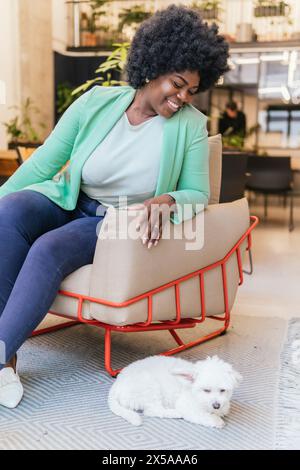 Una donna allegra gode di un momento di relax con un cane bianco seduto in uno spazio di coworking alla moda Foto Stock