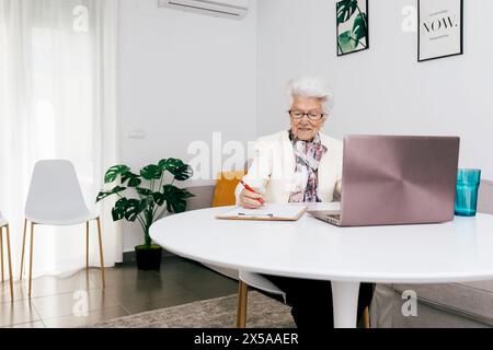 Una donna anziana è concentrata sul lavoro a casa, seduto a un tavolo bianco con un portatile, scrivendo appunti con un interno luminoso Foto Stock