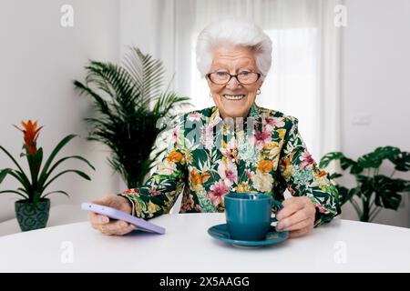 Allegra donna anziana con occhiali e camicia floreale che si gusta una tazza di caffè mentre tiene uno smartphone a un tavolo bianco con piante d'appartamento nel bac Foto Stock