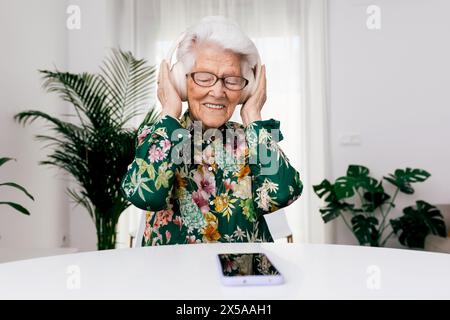 Una donna anziana con una camicia floreale sorride con gioia mentre ascolta la musica con le cuffie nella sua casa luminosa Foto Stock