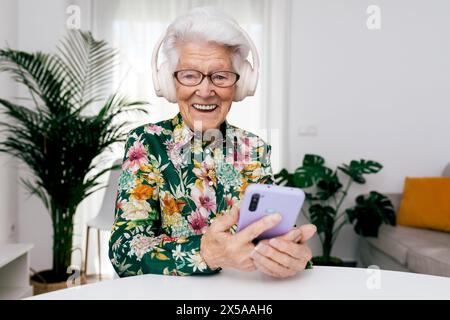 Una donna anziana allegra con i capelli bianchi indossa cuffie di grandi dimensioni e utilizza uno smartphone a casa, il suo sorriso trasuda felicità Foto Stock