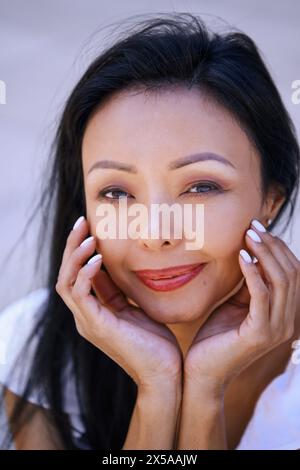 Ritratto ravvicinato di una donna serena che sorride dolcemente con le mani sulle guance, irradiando calma e soddisfazione Foto Stock