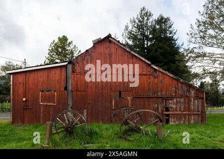 Vecchia, vecchia, rossa, baracca in legno e ruote di carri d'epoca rotte nella California rurale settentrionale. Foto Stock