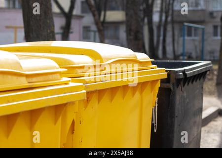 Un contenitore di scarico giallo in materiale composito si trova accanto al contenimento dei rifiuti di un cilindro in plastica nera vicino a una recinzione in legno Foto Stock