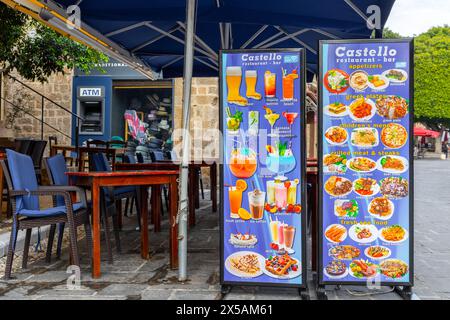 Rodi, Grecia - 12 giugno 2023: Cartelli del menu per cibo e bevande presso il ristorante chiuso con sedie e tavoli. Bancomat in background Foto Stock