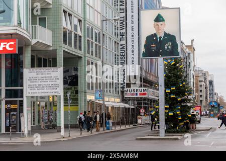 Berlino, Germania - 16 dicembre 2023: Checkpoint charlie con albero di natale e bandiere urbanane. Fotografia del soldato americano e della gente visibile per strada Foto Stock