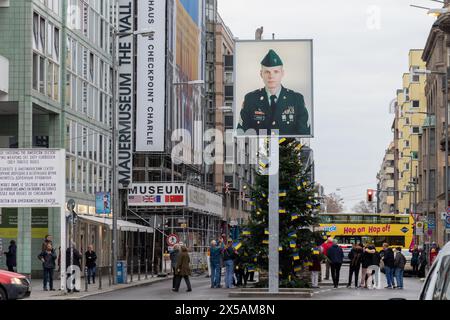 Berlino, Germania - 16 dicembre 2023: Checkpoint charlie con albero di natale e bandiere urbanane. Fotografia del soldato americano e della gente visibile per strada Foto Stock