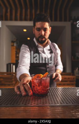 Barman esperto in un gilet che presenta un cocktail negroni appena fatto con una guarnizione di buccia d'arancia Foto Stock