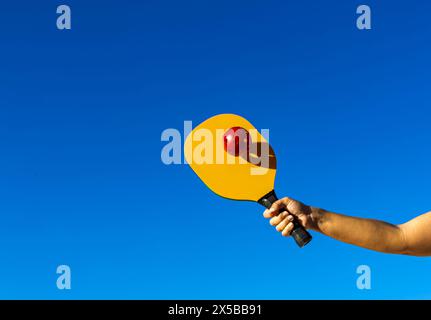 Braccio che regge il picchetto e colpisce la palla. Racchetta da picchetto gialla che colpisce una palla rossa, con il cielo di sfondo e lo spazio di copia Foto Stock