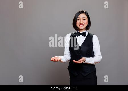 La receptionist del ristorante sorridente donna asiatica accoglie gli ospiti e mostra con ritratti delle mani. La cordiale cameriera in uniforme saluta i clienti mentre posa e guarda la fotocamera Foto Stock