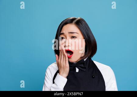 Sonnolenta, giovane aiutante della reception che sbadiglia e copre la bocca con il braccio. La cameriera del ristorante era stanca e lavorava troppo e indossava l'uniforme si sentiva esausta mentre posava su sfondo blu Foto Stock