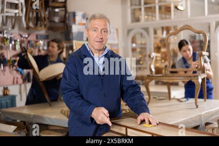 L'uomo anziano esperto restauratore con attenzione e cura ripristina la cassettiera in officina di restauro Foto Stock