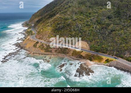 Vista aerea delle onde che si infrangono sotto una strada tortuosa lungo una costa spettacolare vicino a Lorne sulla Great Ocean Road a Victoria, Australia. Foto Stock