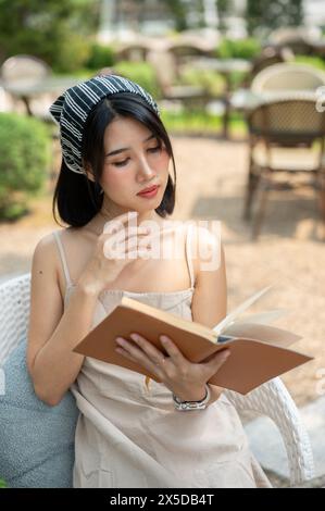 Una bella donna asiatica in un bel vestito si rilassa in un bel giardino in una giornata luminosa, leggendo un libro. tempo libero, hobby, stile di vita Foto Stock