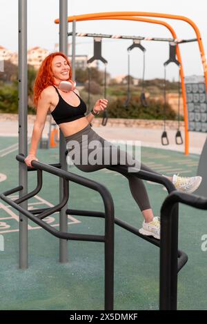 Una donna sorridente che fa jogger in abbigliamento sportivo si rilassa in palestra dopo l'allenamento mattutino. Buon corridore rosso che fa una pausa mentre si siede sull'attrezzatura sportiva nel parco estivo. Foto Stock