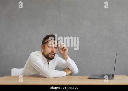 Uomo seduto alla scrivania con un computer portatile e che guarda la fotocamera con un volto che esprime la sua sorpresa Foto Stock