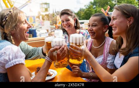 Gruppo di donne in abiti tradizionali tracht brindando con tazze da birra al festival oktoberfest in germania, parco divertimenti sullo sfondo Foto Stock