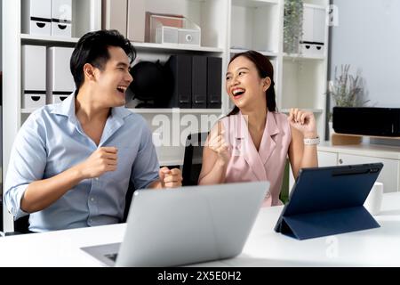 Felicissimo uomo d'affari e donna asiatica sorridendo e ballando con felicità e celebrando il successo mentre si è seduti sulla scrivania con computer portatile e tablet in ufficio. - Un posto di lavoro di successo e divertente. Foto Stock