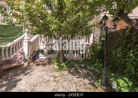 un patio con pavimenti di ghiaia bianca con balaustra di cemento bianco su scale e terrazza, molte piante, una lanterna e alcuni alberi Foto Stock