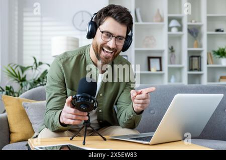 Allegro podcaster maschile in cuffia che registra un podcast dal vivo. Sta gestendo e sorridendo calorosamente nella sua comoda sistemazione a casa. Foto Stock