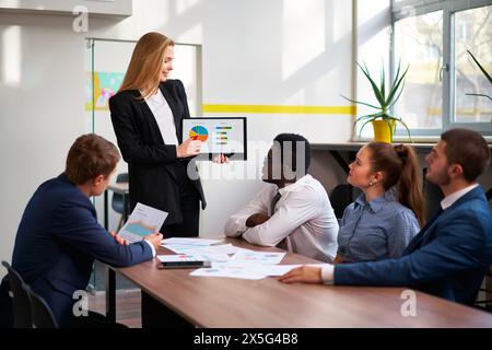 Una donna leader presenta un grafico a torta colorato sul tablet ai colleghi durante la riunione d'ufficio. Un team attento si occupa di pianificazione strategica e analisi Foto Stock