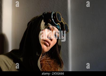 n coinvolgente ritratto di una donna con cappuccio che indossa occhiali steampunk, il suo volto illuminato da un ardito flusso di luce solare, creando un gioco dinamico di luce e. Foto Stock