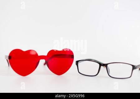 Un paio di occhiali da sole rossi a forma di cuore e un paio di occhiali neri sono seduti su un tavolo bianco. Concetto di giocosità e fantasia Foto Stock