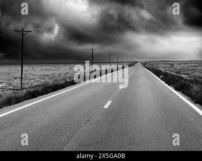 Un paesaggio bianco e nero che mostra una strada di campagna a due corsie vuota che si estende in lontananza con nuvole di tempesta in avvicinamento. Foto Stock