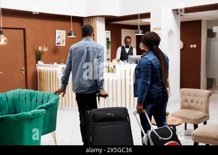 Vista posteriore della giovane coppia di famiglie afroamericane in arrivo all'hotel del resort, persone che portano i bagagli alla reception per il check-in. Concetto di persone, viaggi e vacanze Foto Stock