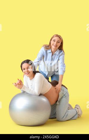 Doula matura e donna incinta si allenano con fitball su sfondo giallo Foto Stock