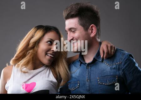 Il giovane con i capelli corti e la camicia in denim abbraccia delicatamente una donna con lunghi capelli biondi evidenziati, entrambi sorridenti Foto Stock