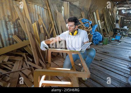 Uomo con occhiali di sicurezza, protezioni per l'udito e guanti impegnato nella lavorazione del legno. Misurare un pezzo di una sedia, con un metro a nastro tenendo in mano un di Foto Stock