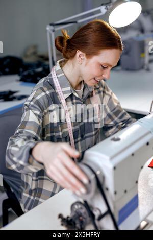 giovane sarta caucasica nella cucitura in fabbrica con la moderna macchina da cucire industriale, rossa lavorata femminile piacevole e accurata in officina, sartoria. concetto di produzione di abbigliamento Foto Stock