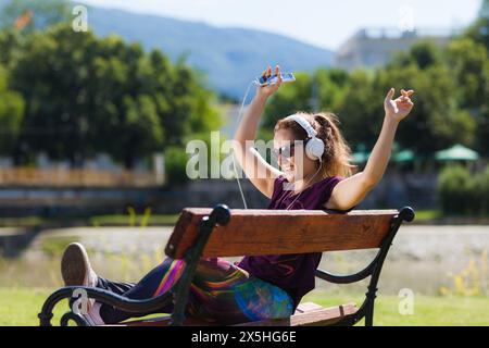 Una giovane donna siede su una panchina del parco, ascoltando con gioia la musica con le cuffie bianche, facendo un segno di pace, in una giornata di sole con uno sfondo naturale. Foto Stock