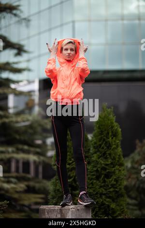 Una giovane donna sta su un piedistallo in città, regolando il cappuccio della sua giacca sportiva arancione brillante, trasmettendo un senso di fitness e stile urbano. Foto Stock