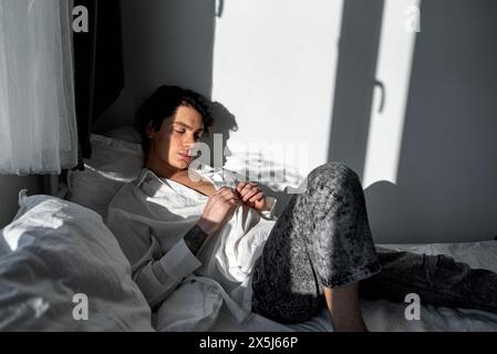 il bell'uomo assonnato si siede sul letto e si prepara a lasciare la camera d'albergo, indossa il suo costume da lavoro. passione per il suo amante e sdraiato sul letto, vista laterale ravvicinata Foto Stock