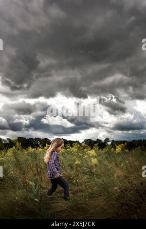 Giovane ragazza con lunghi ricci che cammina attraverso fiori gialli cielo nuvoloso Foto Stock