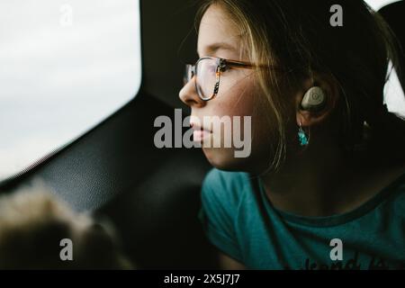 Tween girl con gli occhiali guarda fuori dal finestrino dell'auto durante il viaggio in auto con le cuffie Foto Stock