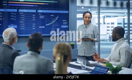 Ufficio moderno diversificato: L'imprenditrice digitale femminile nera di successo utilizza lo schermo TV con Big Data, statistiche, parla della crescita aziendale, discute della strategia con investitori, top manager, dirigenti Foto Stock