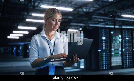 Tecnico IT del data center donna in piedi presso il corridoio del rack server con computer portatile. Sta ispezionando visivamente qualcosa mentre guarda lo schermo. Concetto di ufficio notturno Foto Stock