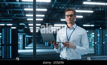 Uomo d'affari creativo in piedi in un ufficio moderno, tenere il computer portatile e guardare la fotocamera. Managing Director che pianifica progetti di lavoro presso la sala di lavoro serale Foto Stock