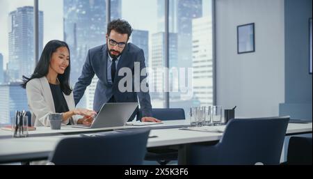 Modern Office: CEO aziendale di successo seduto alla sua scrivania lavorando su laptop e Consulting Top Manager. Gli uomini d'affari sorridenti discutono degli investimenti nell'avvio dell'e-Commerce digitale. Foto Stock