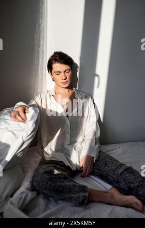 Bell'uomo in pigiama con gli occhi chiusi in camicia bianca e pantaloni si siede sul letto a casa, foto ravvicinata. buon giorno. Foto Stock