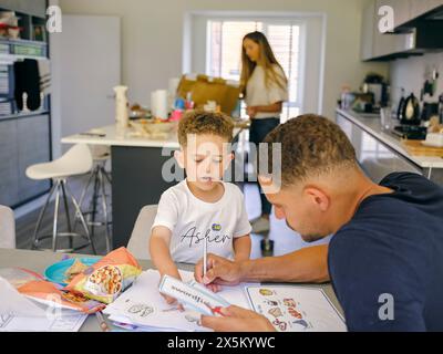 Padre che assiste il figlio a fare i compiti a casa, madre in background Foto Stock