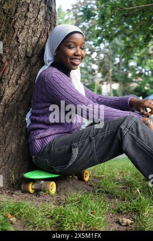 Regno Unito, Londra, giovane donna che indossa un hijab seduto sullo skateboard accanto al tronco dell'albero Foto Stock