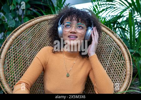 Donna sorridente con cuffie sedute in una poltrona in rattan in stile tropicale Foto Stock